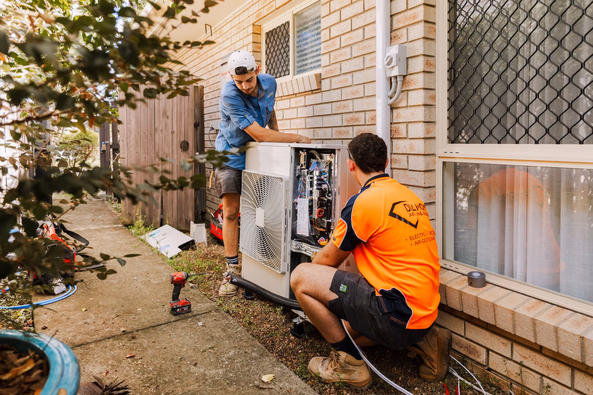 Two technicians working on an outdoor air conditioning unit beside a house wall and window.
