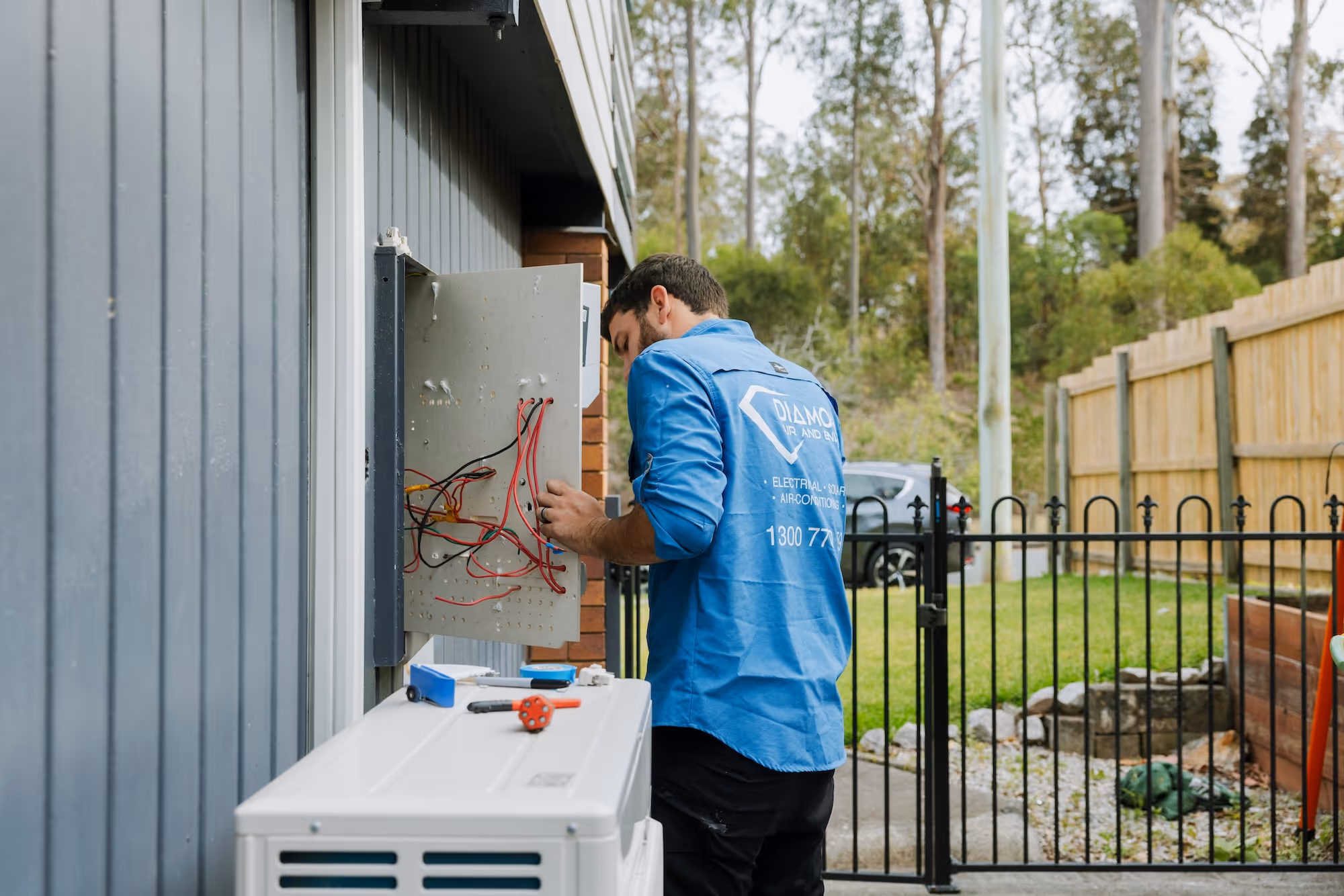 Technician in blue shirt working on electrical wiring in an outdoor metal box on a building wall.