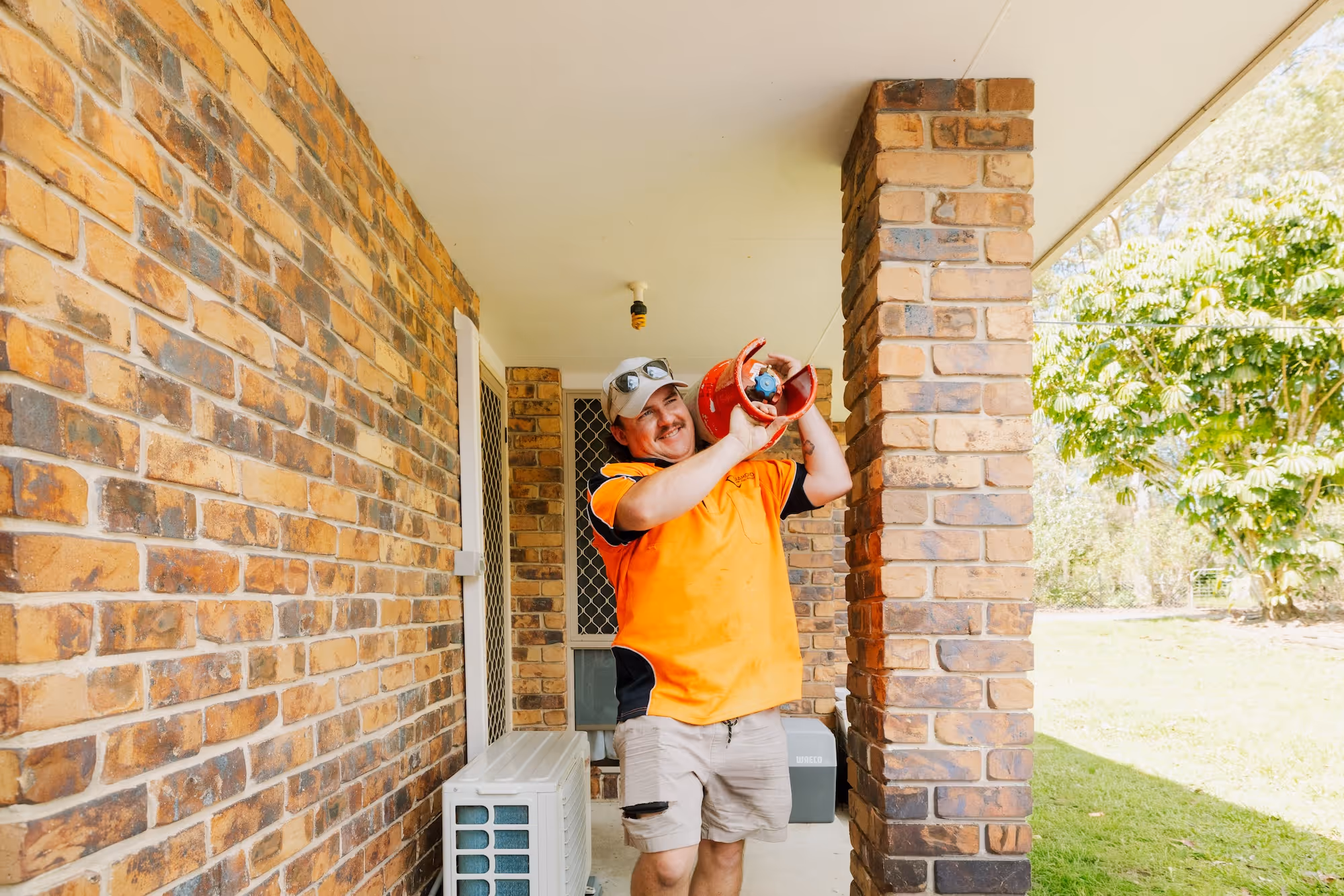 Man in orange shirt and cap carrying a red gas cylinder on a brick porch.