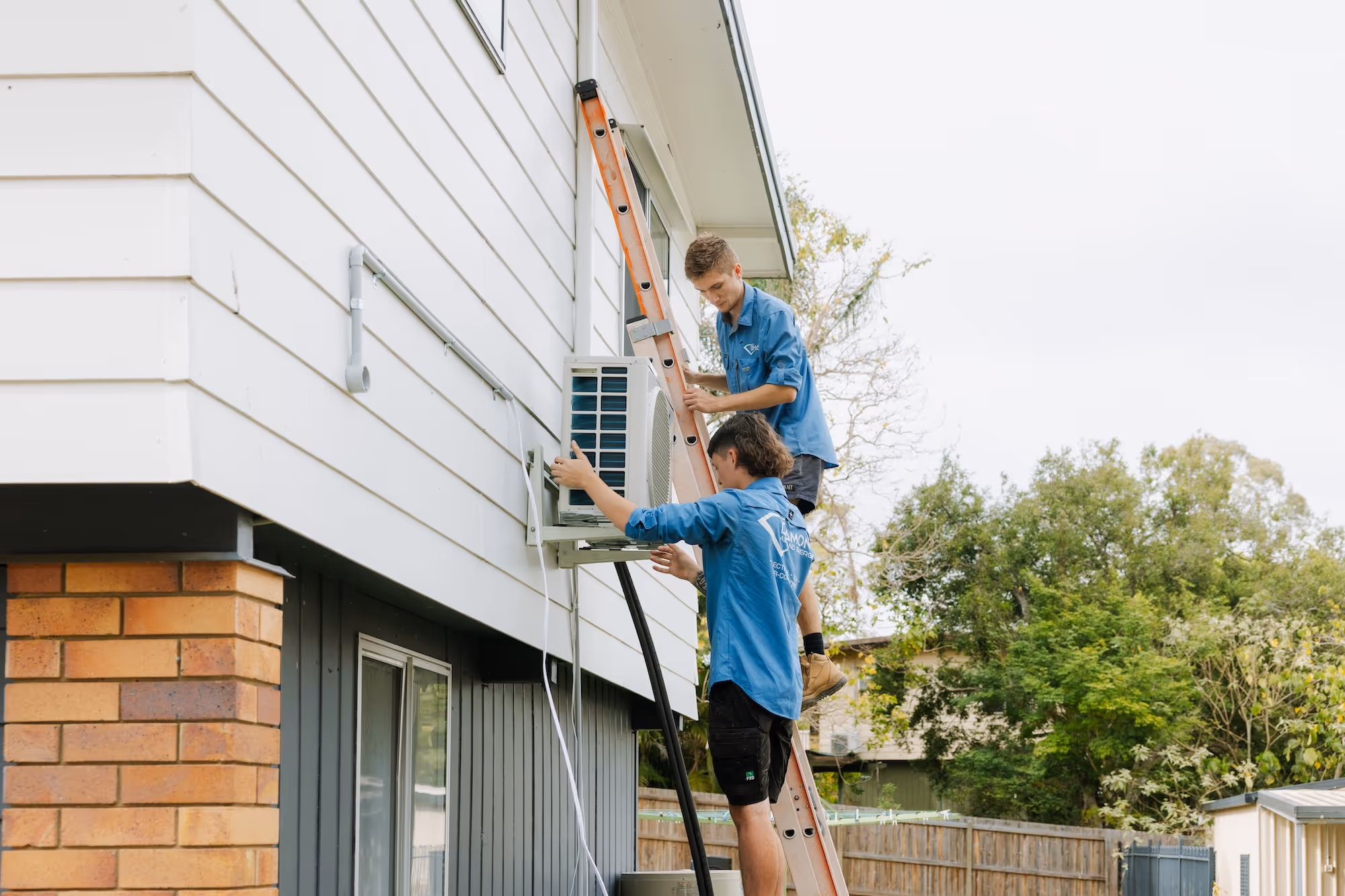 Two workers in blue shirts installing an outdoor air conditioning unit on a white house wall using a ladder.