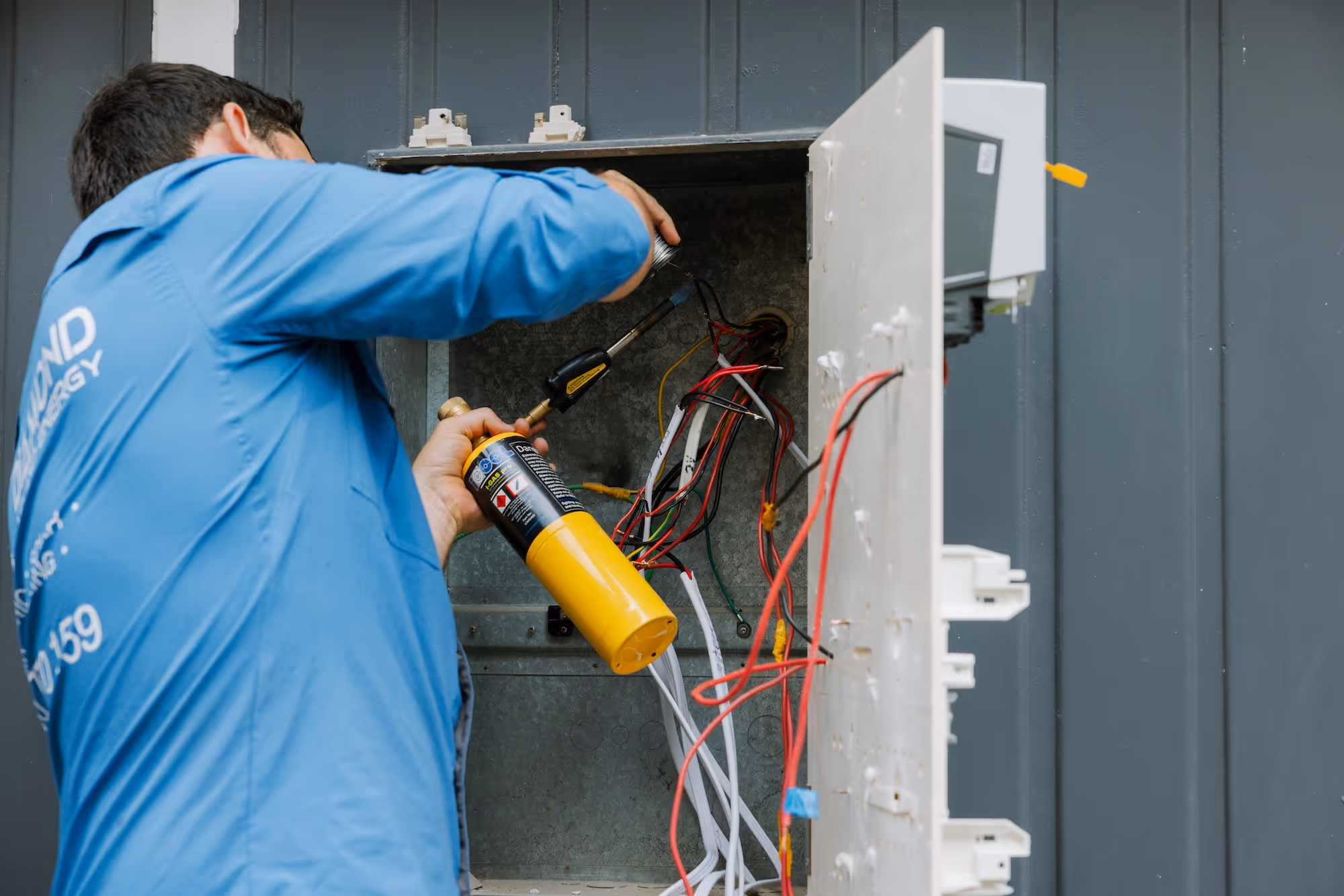 Technician in a blue shirt working on a fuse box with a yellow torch and wires.