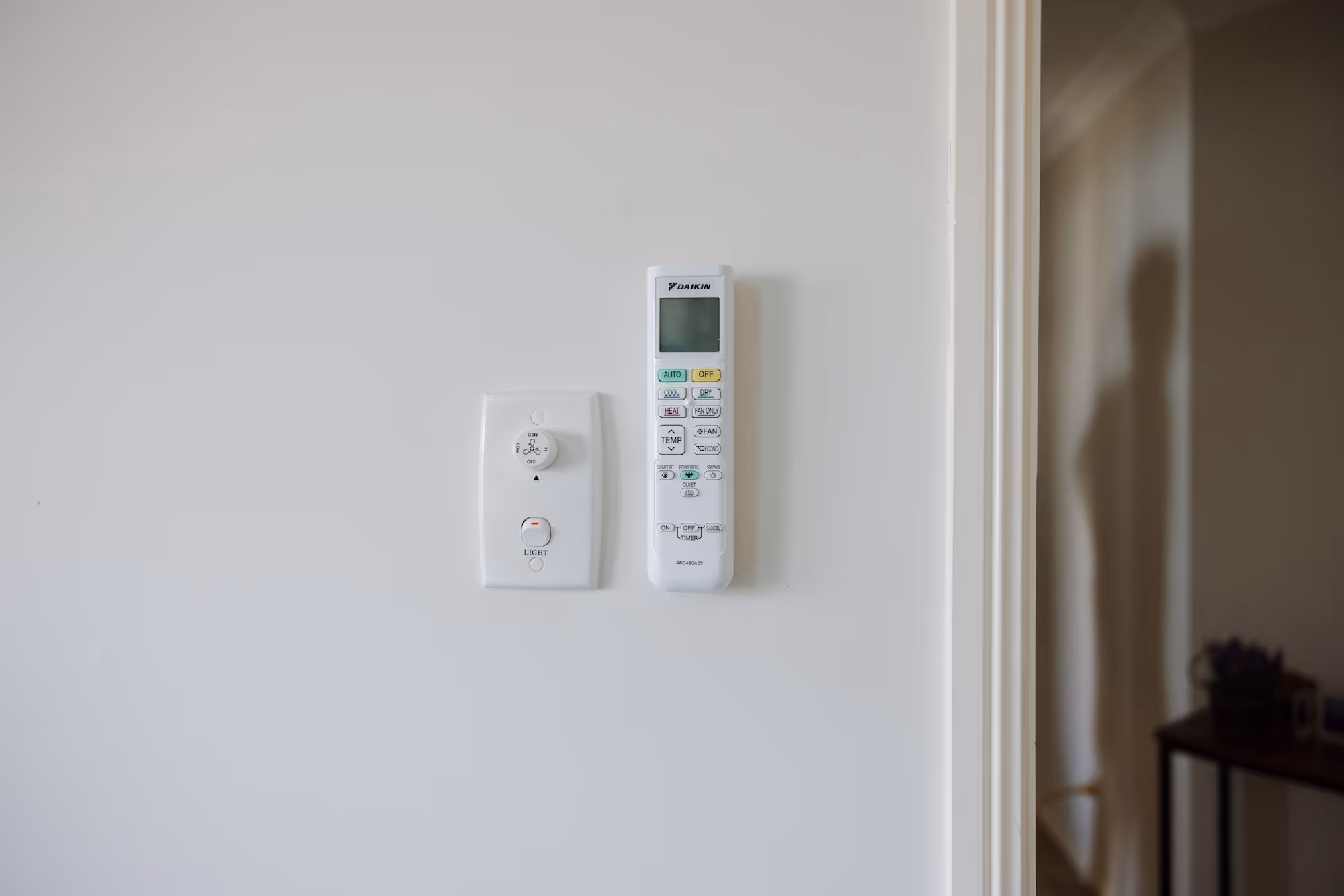 White wall with a Daikin air conditioner remote and a fan speed dial switch with a light button.