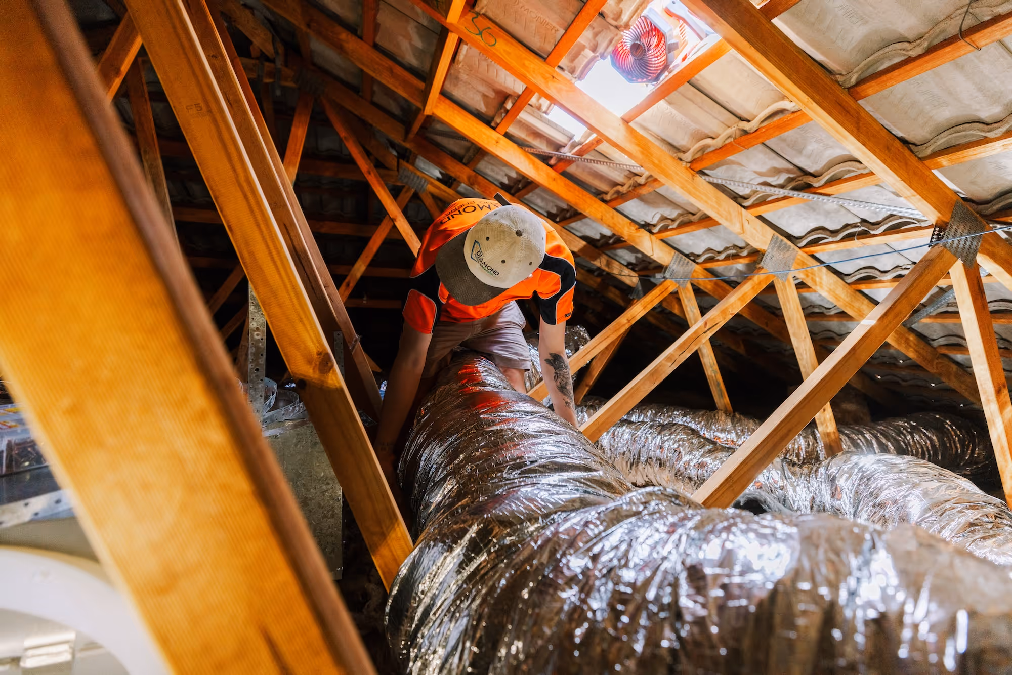 Worker adjusts large silver ducting in wooden attic with visible roof structures and insulation.
