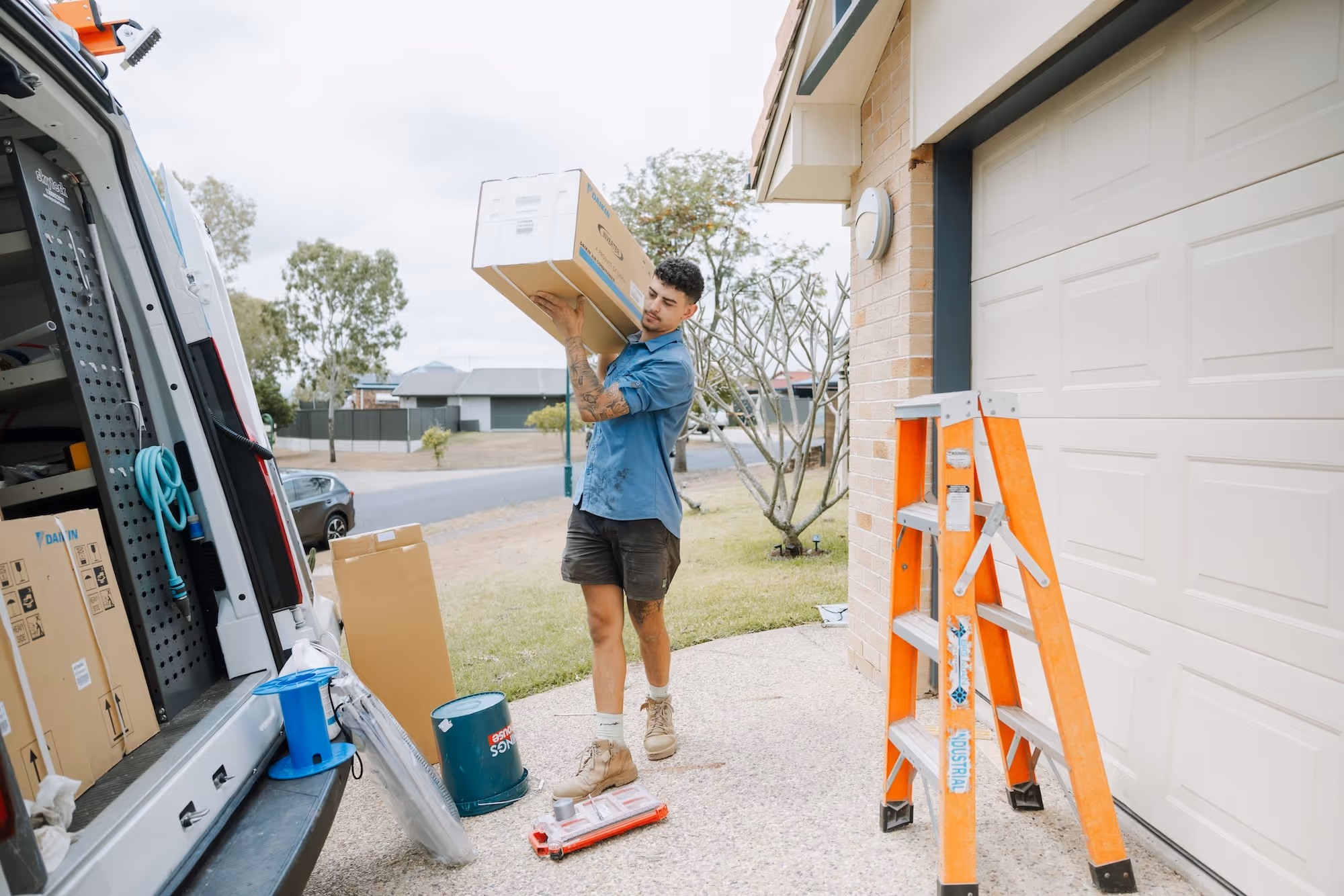 Man carrying a large boxed air conditioning unit from a work van near a house garage with ladder.