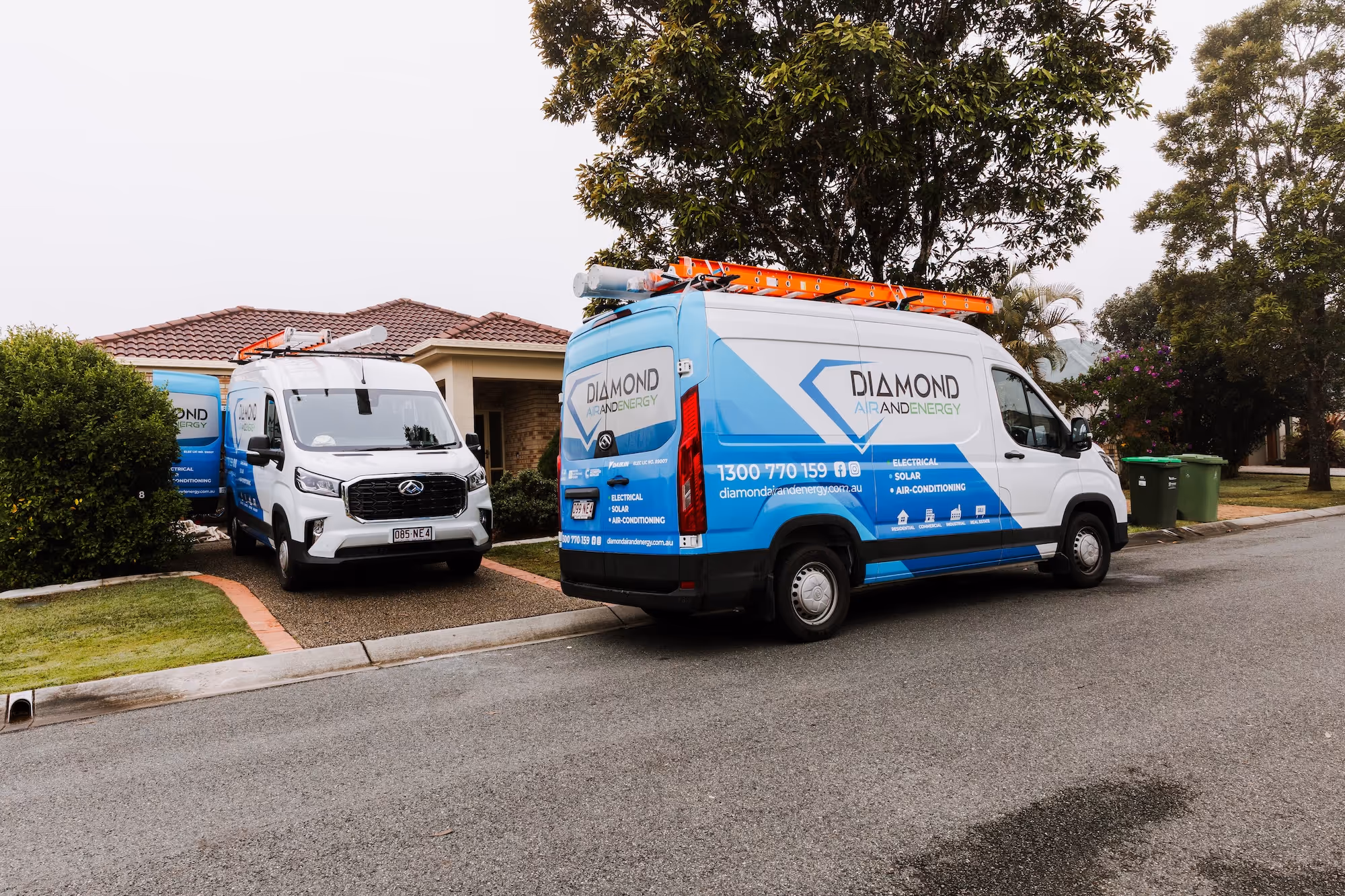Two Diamond Air and Energy vans with ladders parked outside a suburban house on a street.