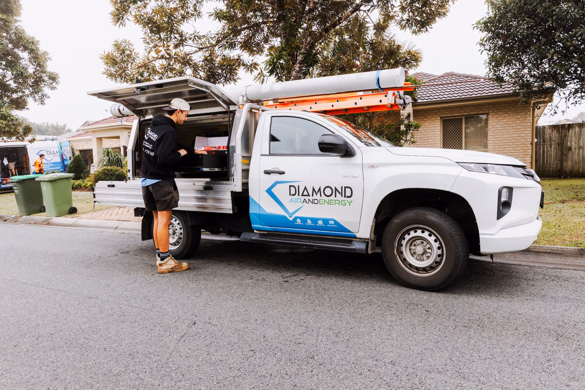 Worker in black hoodie next to white Diamond Air and Energy truck parked on suburban street.
