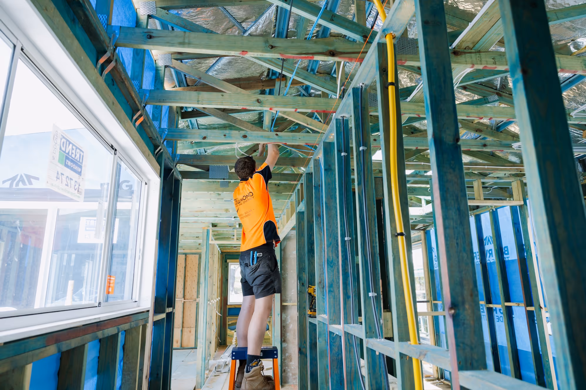 Construction worker in an orange shirt installing wiring in a wooden frame building under construction.
