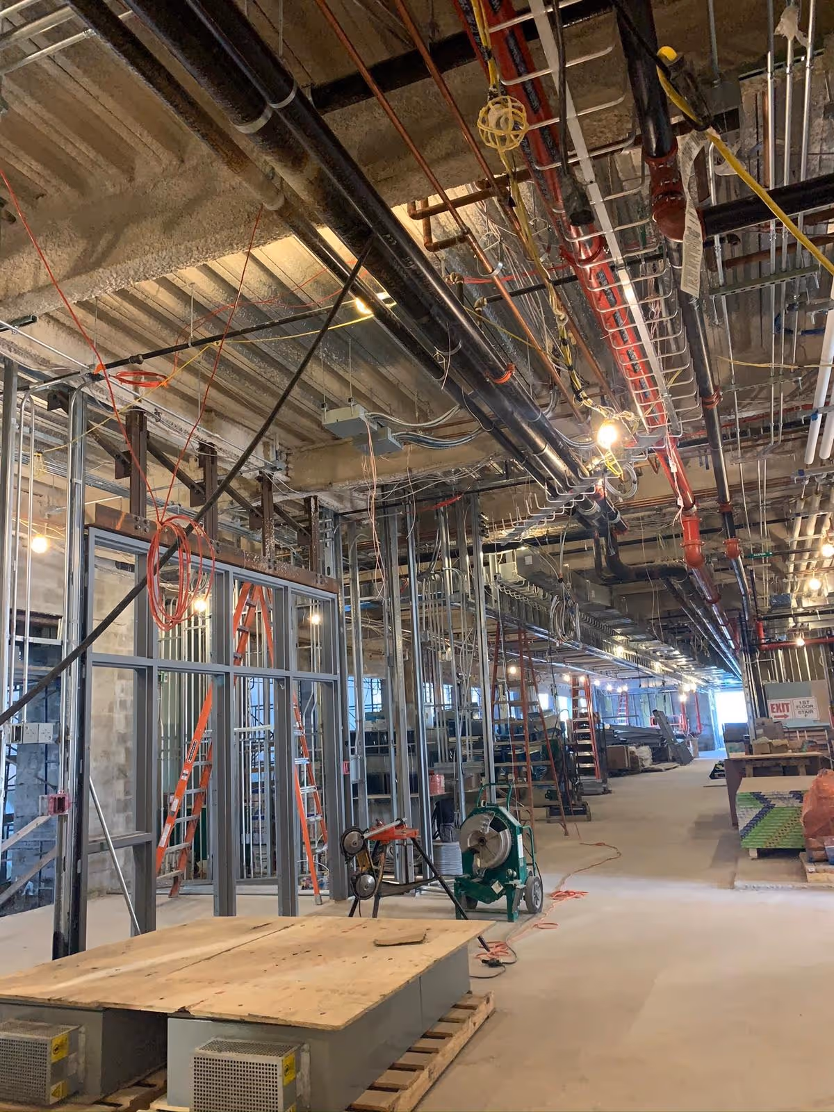 Interior view of an active institutional electrical upgrade showing exposed metal framing, overhead electrical conduits, piping, and commercial wiring systems.