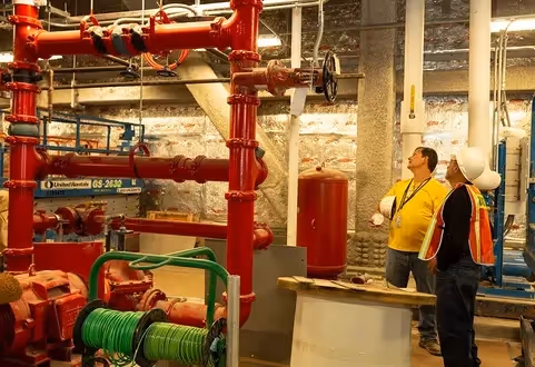 Commercial building basement interior detailing mission-critical public address systems, red fire alarm piping, and electrical conduit lines with two technicians reviewing infrastructure.