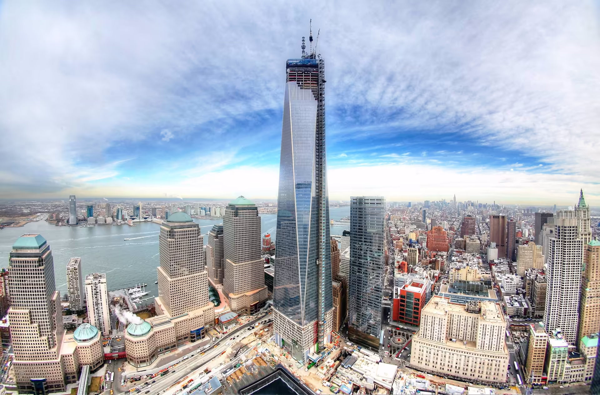 Wide-angle aerial view of the One World Trade Center skyscraper and surrounding lower Manhattan skyline in New York City, overlooking the Hudson River under a bright blue sky with clouds.