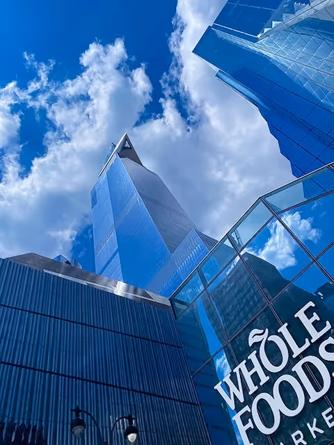 Low-angle view looking up at modern glass skyscrapers in New York City under a cloudy blue sky, with the glass facade of a Whole Foods Market reflecting the clouds in the foreground.