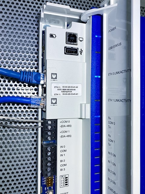Close-up of a wall-mounted building management system (BMS) network control module with blue Ethernet cables plugged in and terminal block wiring.