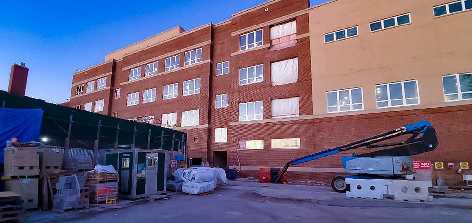 Active construction site of a brick municipal school building with a blue boom lift crane, construction materials, and a temporary site office in the foreground.