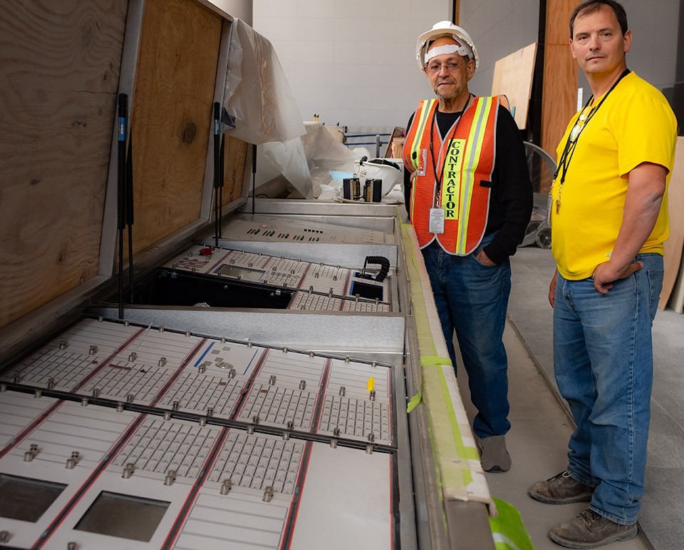 Two electrical contractors in hard hats and high-visibility vests standing next to an open heavy-duty electrical command console and switchboard at the World Trade Center.