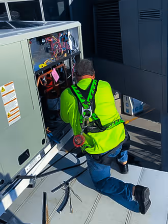 An electrical technician in a neon green shirt and safety harness kneeling on a roof while servicing the open wiring panel of a commercial HVAC unit.