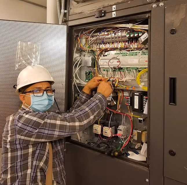 An electrical engineer in a hard hat, face mask, and glasses making adjustments to the wiring inside an open building management system control panel.