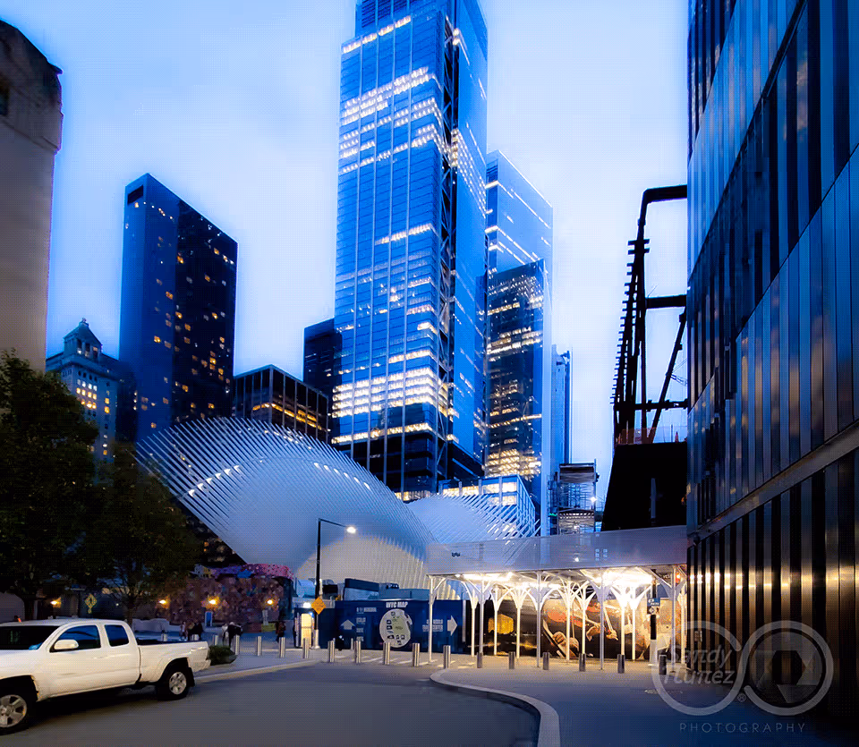 Dusk street-level shot of the World Trade Center Oculus transportation hub lit up at night, framed by modern glass skyscrapers in Lower Manhattan.