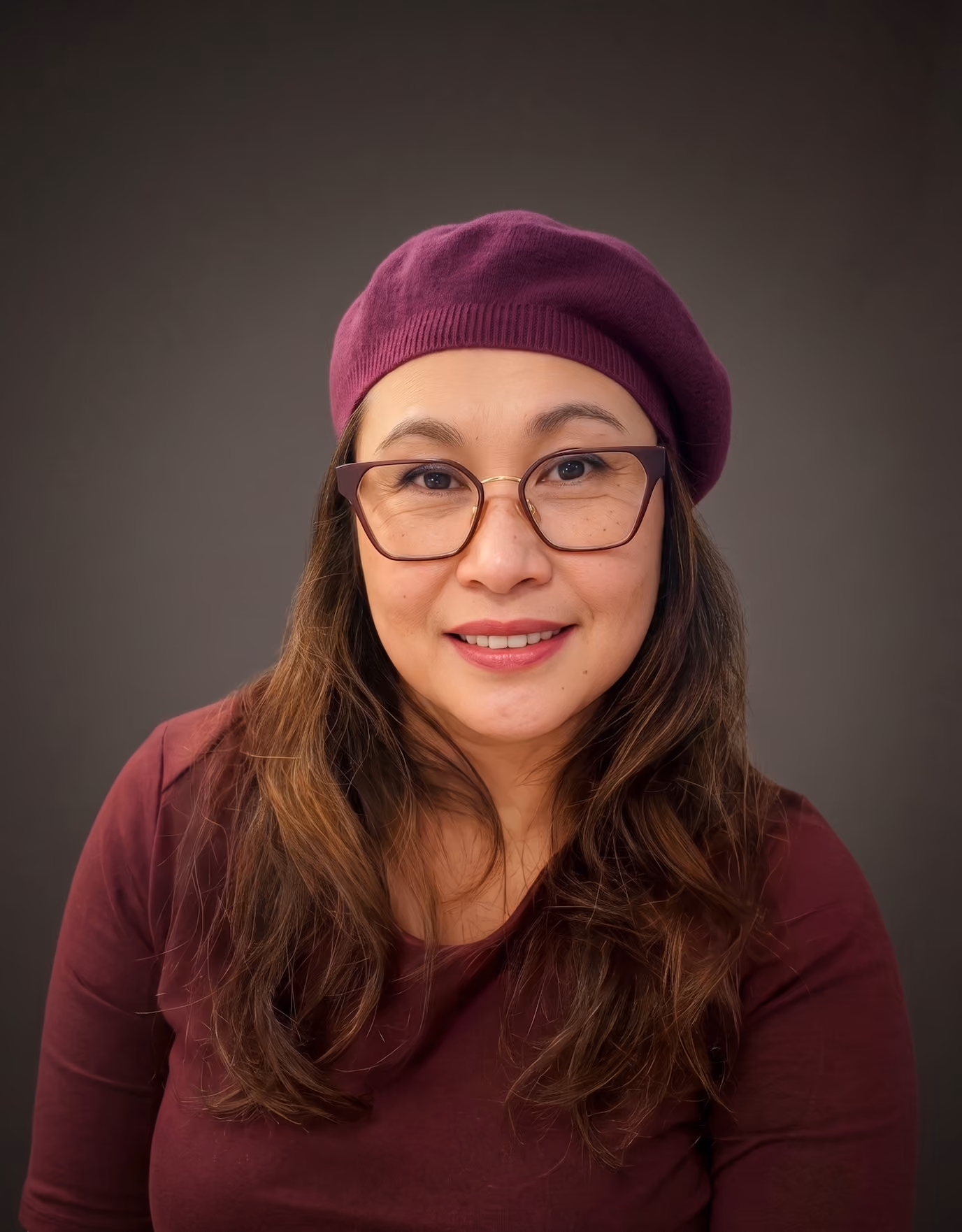 A professional mid-shot corporate portrait of a female founder against a solid, dark gray studio background. The woman is smiling warmly at the camera, wearing her natural long brown hair styled in soft, smooth waves. She wears stylish, thin-framed glasses, a deep maroon top, and a matching plum-colored beret. The lighting is soft and even, highlighting her confident expression.