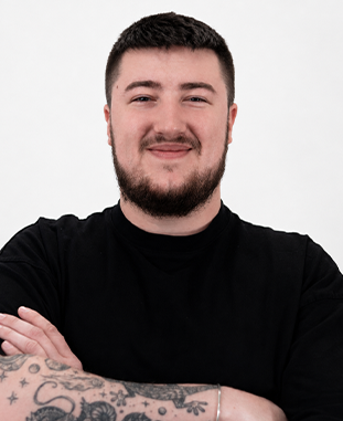 Uśmiechnięty mężczyzna z krótkimi brązowymi włosami i brodą, ubrany w szarą koszulkę.