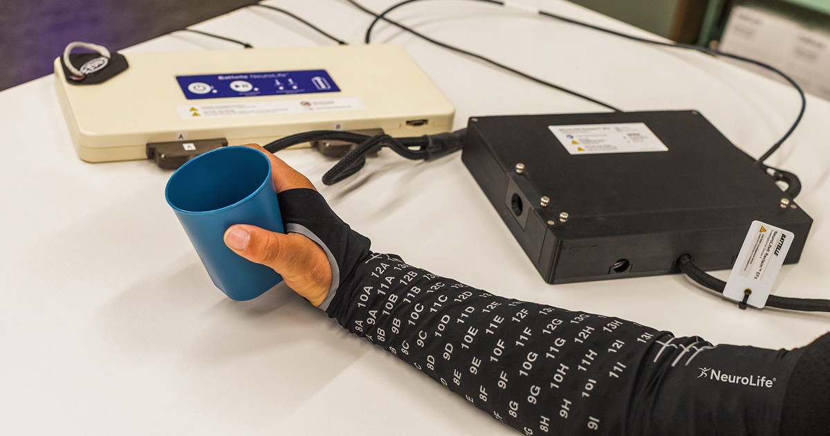 Person wearing NeuroLife sleeve on arm holding a blue cup, connected to medical devices on a table.