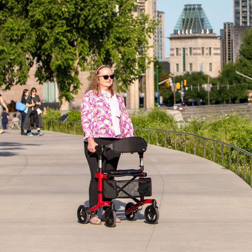Woman using a Simple Comforts rollator walker on an urban path with city buildings in the background.