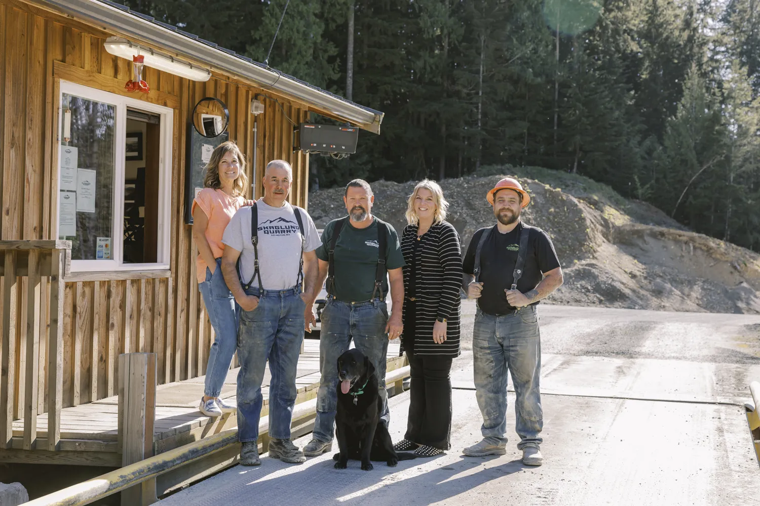 Five quarry workers standing outside a wooden building with a black dog in front, surrounded by a gravel quarry and forest.