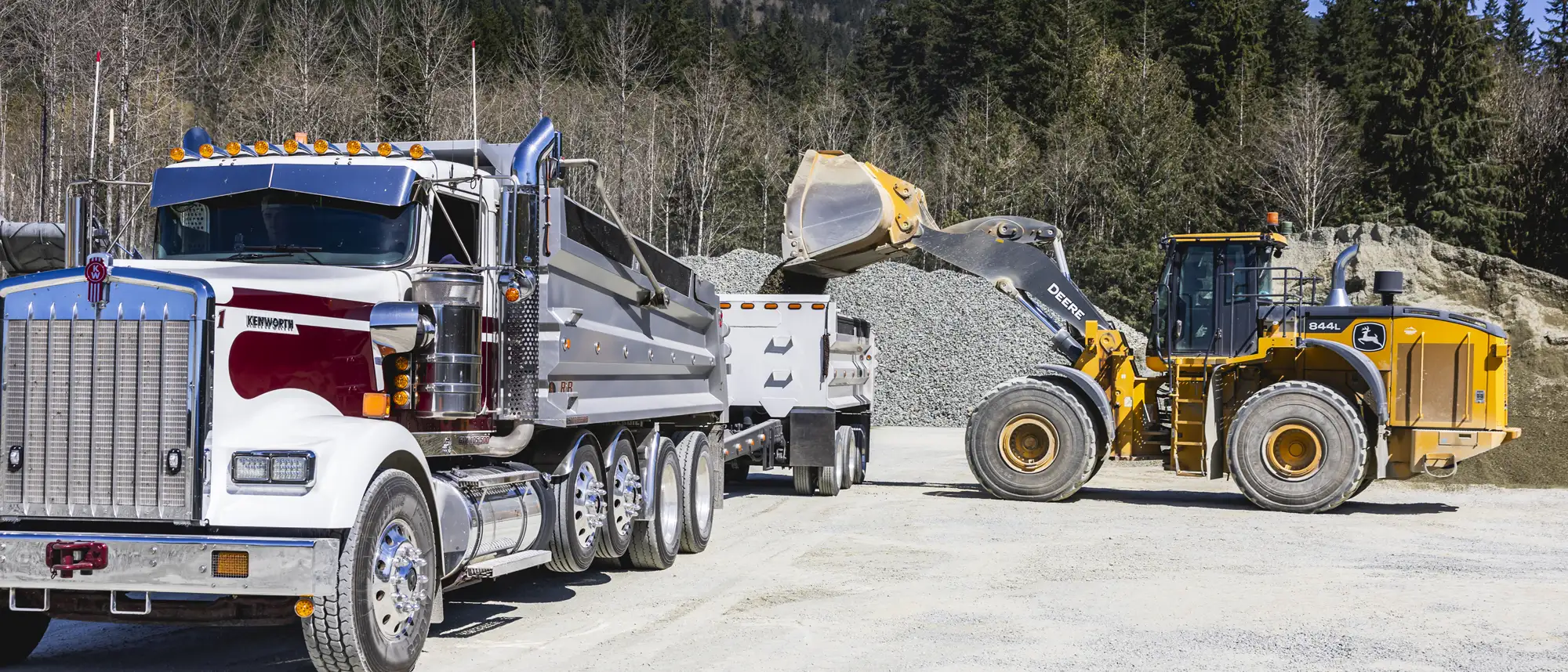Yellow Deere 844L front loader loading gravel into a white and maroon Kenworth dump truck at a quarry.
