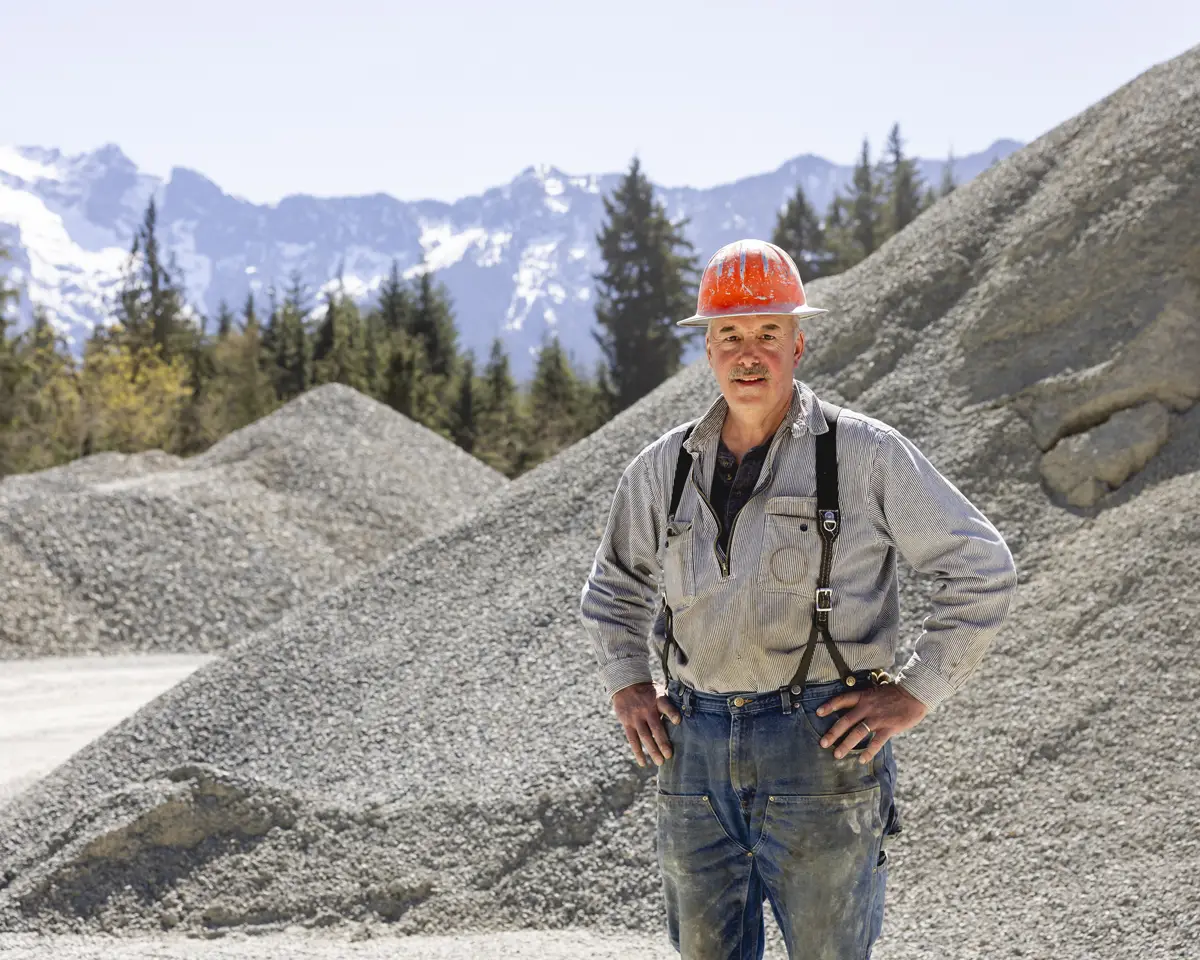 Construction worker wearing an orange hard hat and suspenders standing in front of large gravel piles with a mountainous forest background.