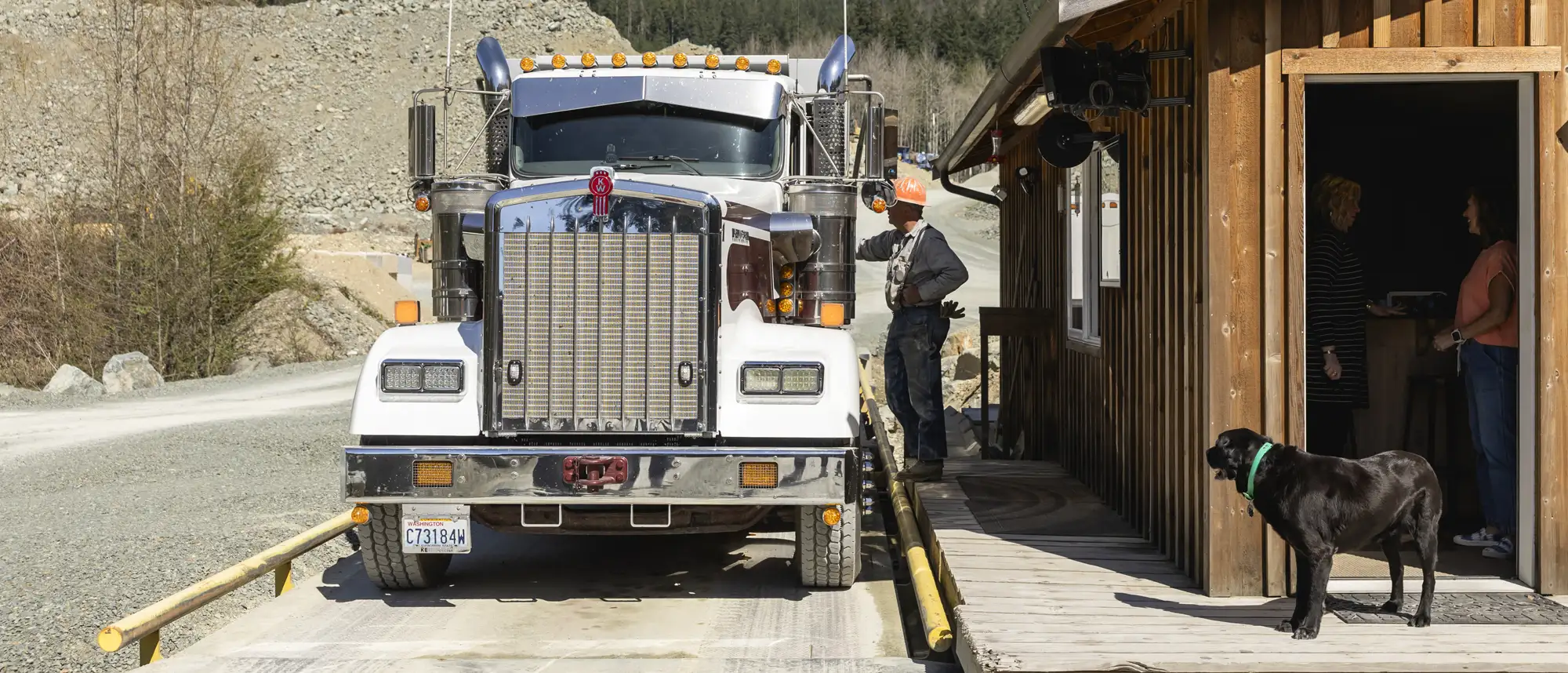 Front view of a large white semi-truck next to a wooden building with a man in a hard hat interacting with the truck and a black dog standing by the building entrance where two women are talking inside.