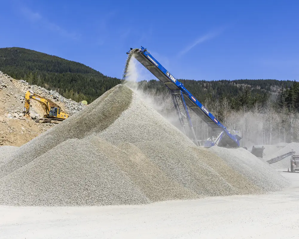 Large pile of gravel being deposited by a blue conveyor belt machine with a yellow excavator and forested hills in the background under a clear blue sky.