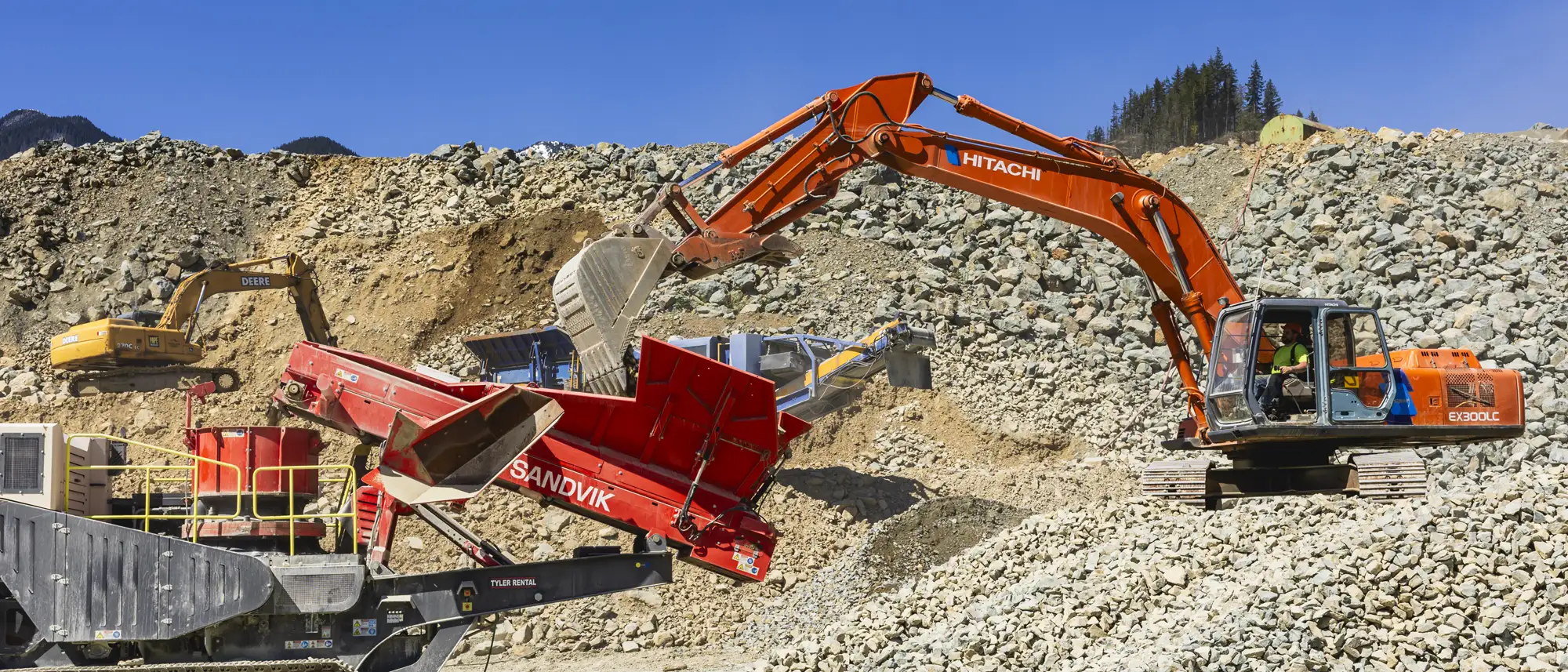 Orange Hitachi excavator loading rocks into a red Sandvik crusher at a rocky quarry site under a clear blue sky.