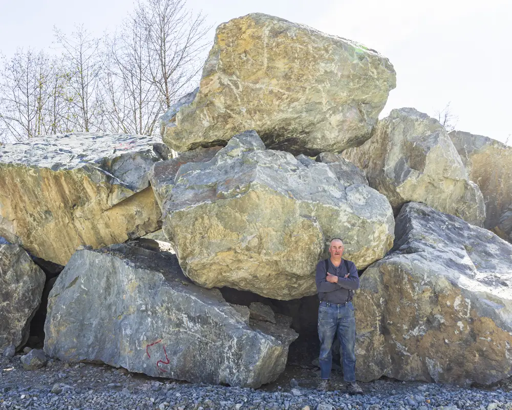 Jetty stone - Man standing with arms crossed in front of large stacked boulders at a quarry with bare trees in the background.