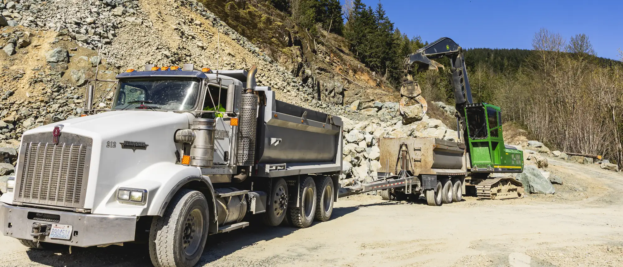 White Kenworth dump truck and green excavator working together to load rocks at a quarry site with rocky hills and trees in the background.