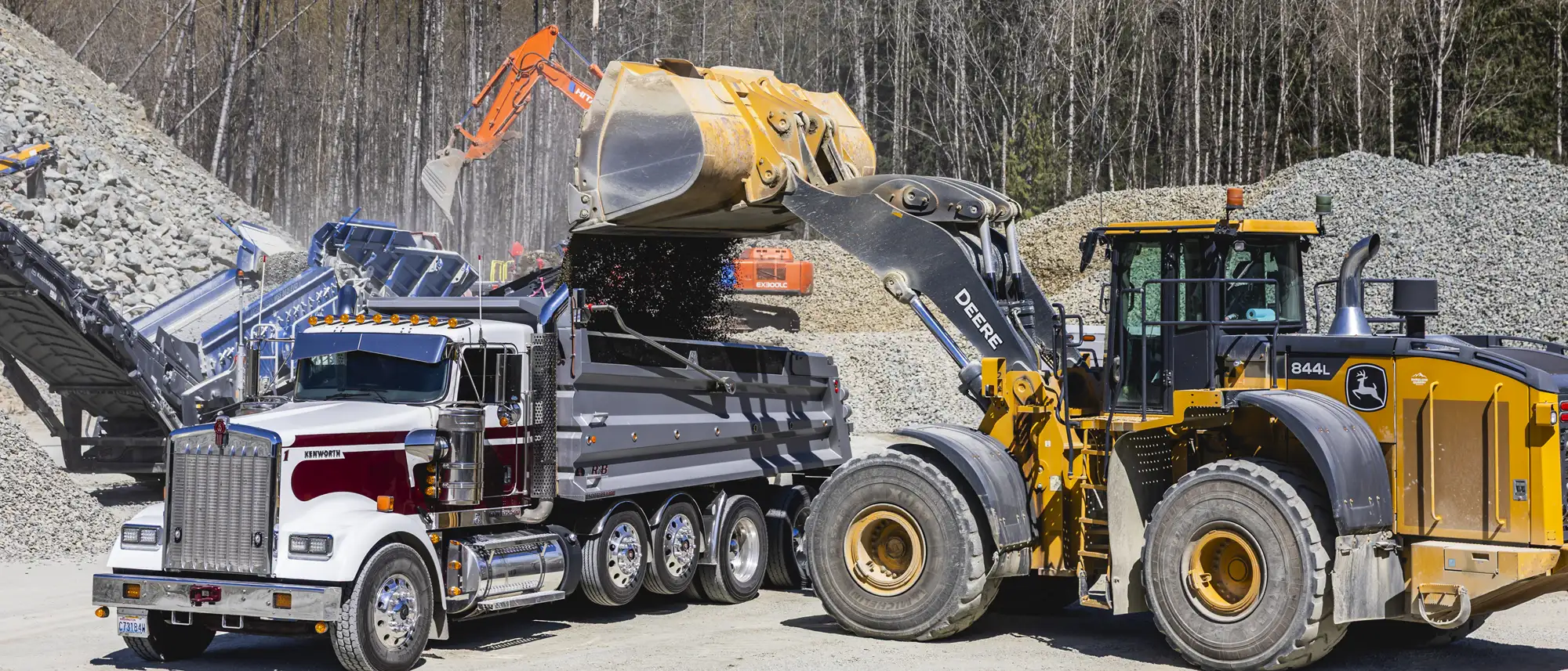 Yellow John Deere 844L loader dumping gravel into white and maroon Kenworth dump truck at a quarry site with gravel piles and machinery in background.
