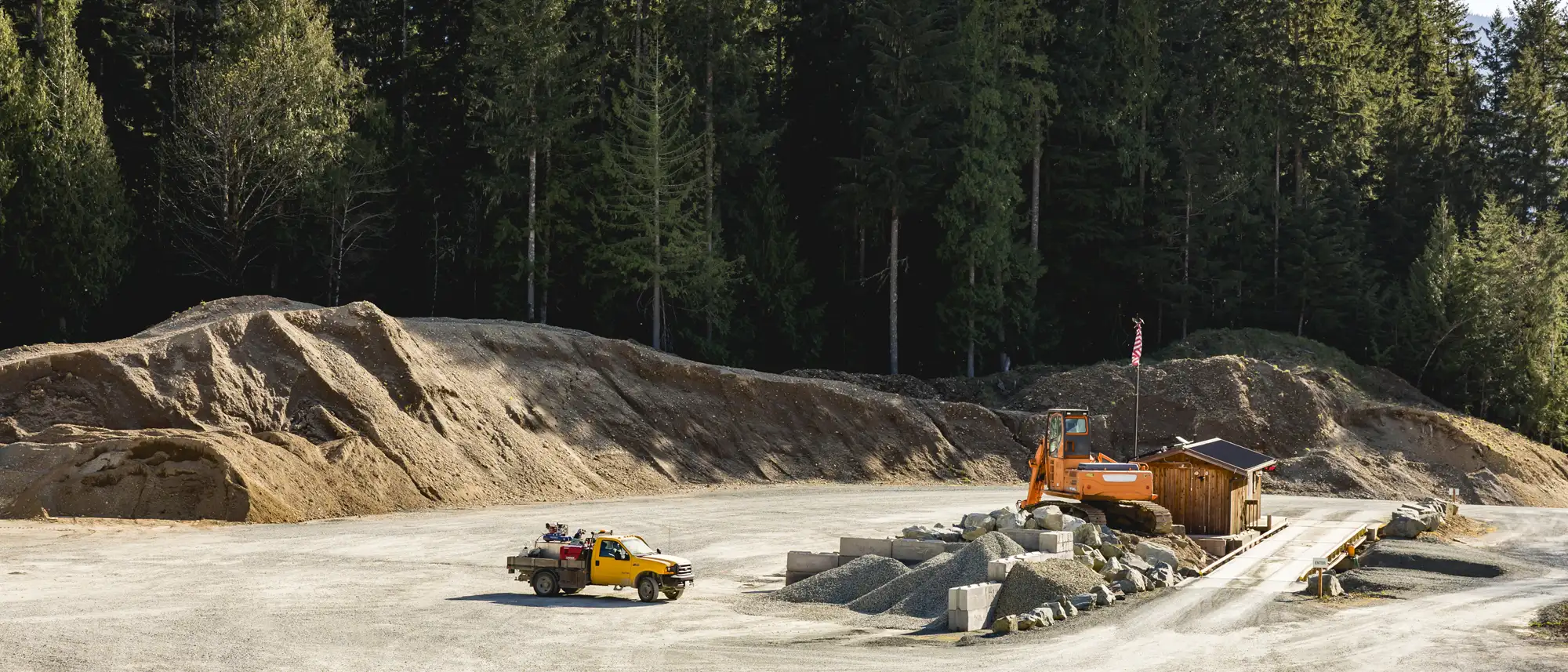 Gravel quarry site with piles of gravel and dirt, a yellow pickup truck, and an orange excavator near a small wooden building.