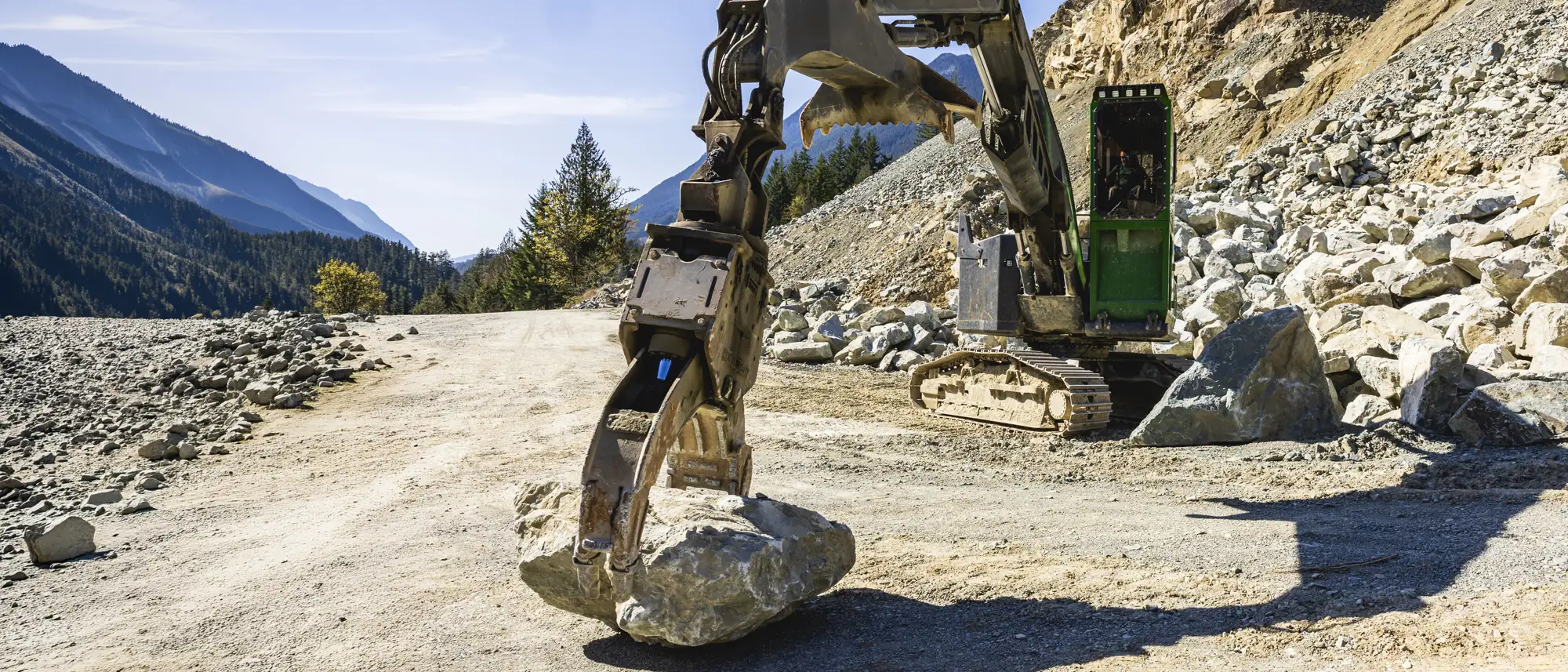 Excavator with a hydraulic rock breaker lifting a large boulder at a quarry site surrounded by mountains.