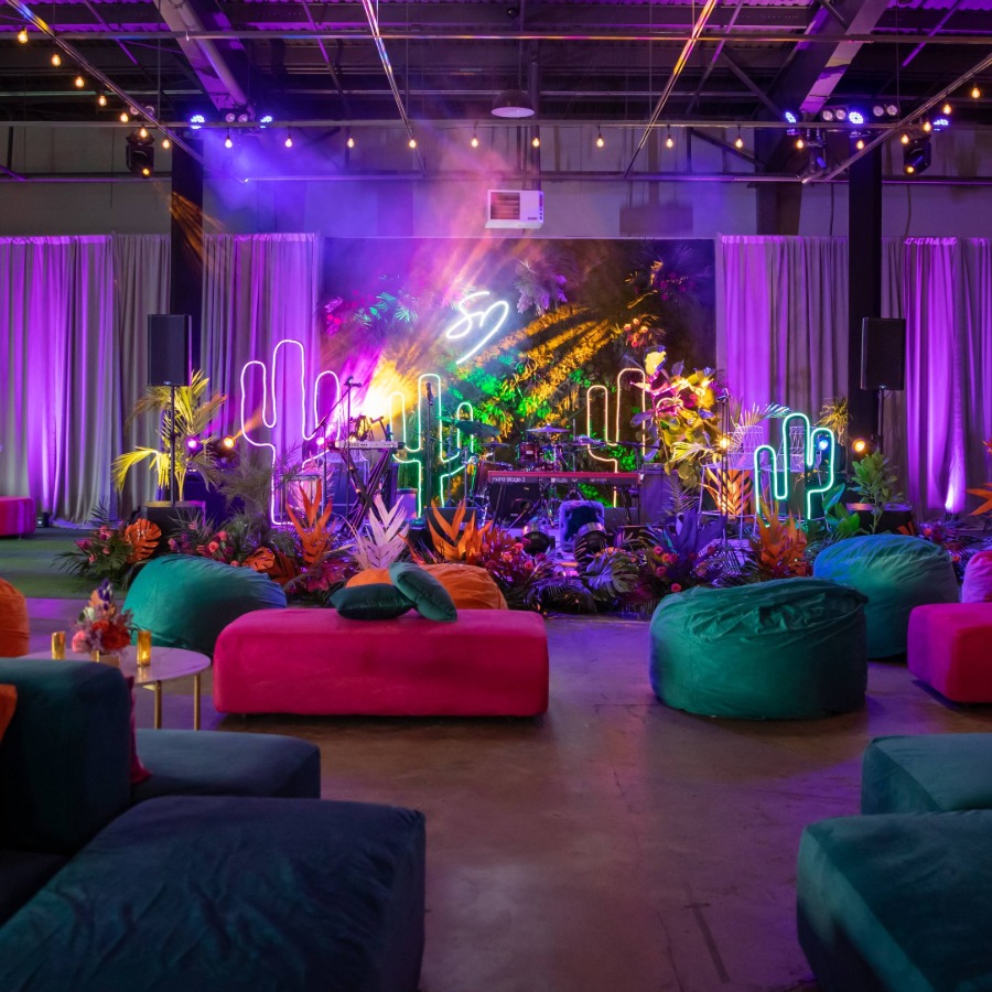 Indoor party setup with neon cactus lights, colorful seating, stage, and purple ambient lighting.