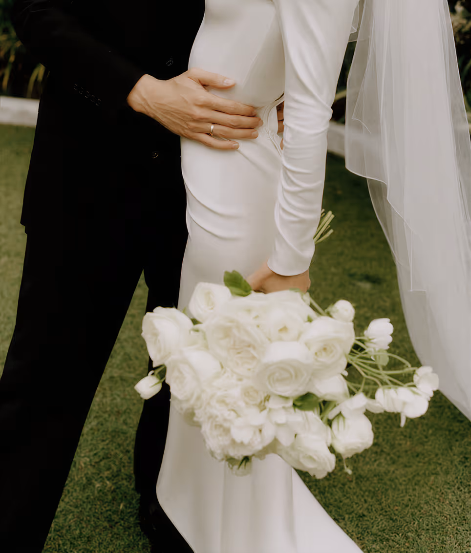 Close-up of a groom in a black suit embracing a bride in a white wedding dress holding a bouquet of white roses and flowers.