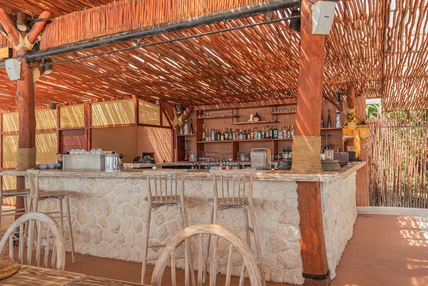 Outdoor bar with stone counter, wooden stools, and a rustic wooden roof with beams and bottles on shelves.