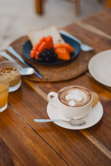 Cup of hot chocolate with latte art on a white saucer and spoon, next to a plate of toast and fruit on a wooden table.