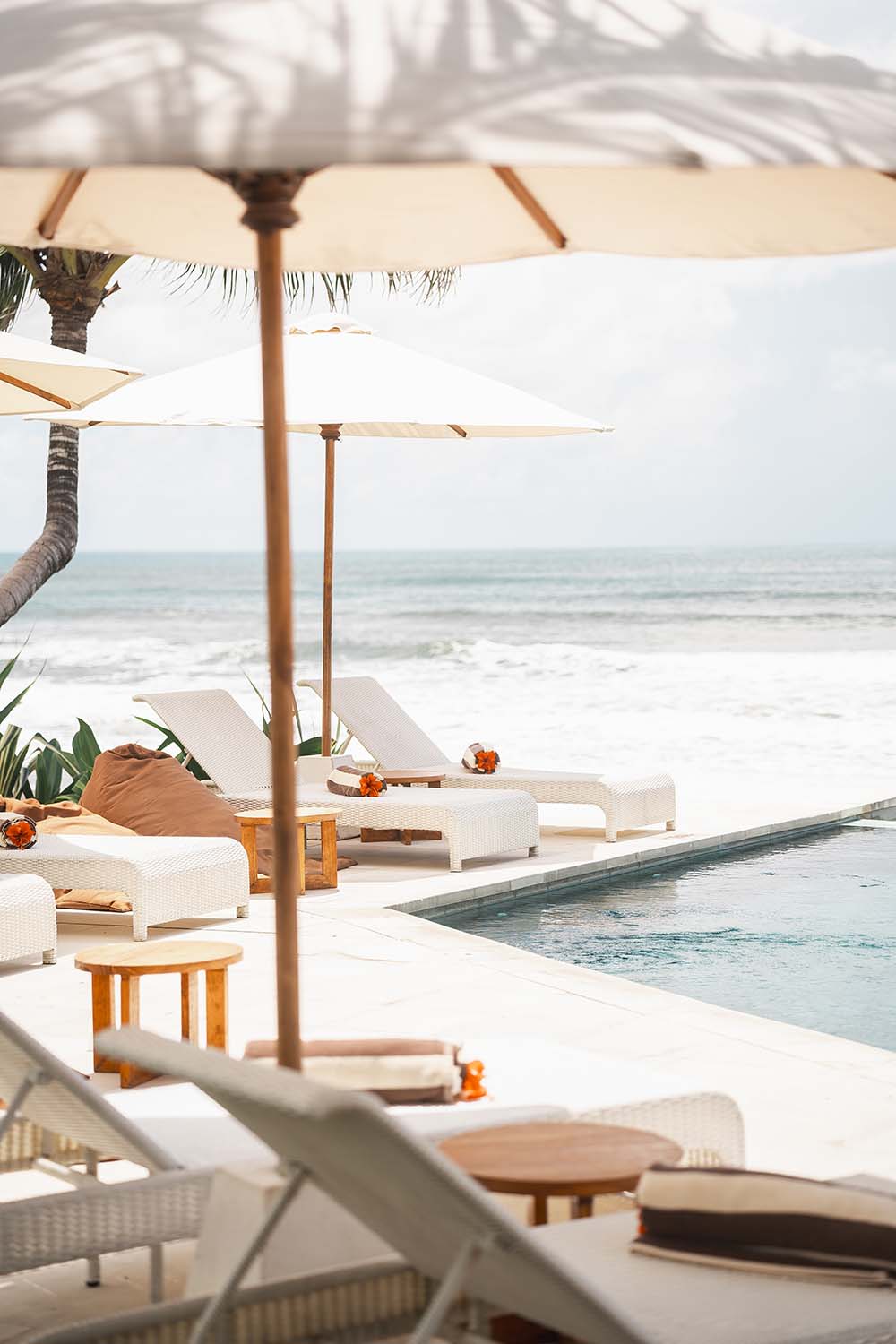 Poolside lounge chairs with umbrellas next to an ocean view and palm trees on a sunny day.