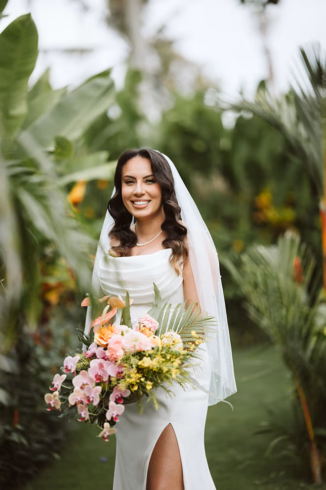 Smiling bride in white dress holding a colorful bouquet in a lush green outdoor setting.