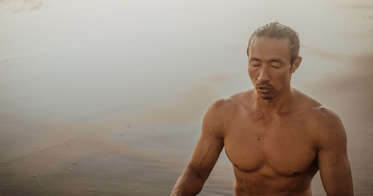 Shirtless man with eyes closed meditating near calm water.