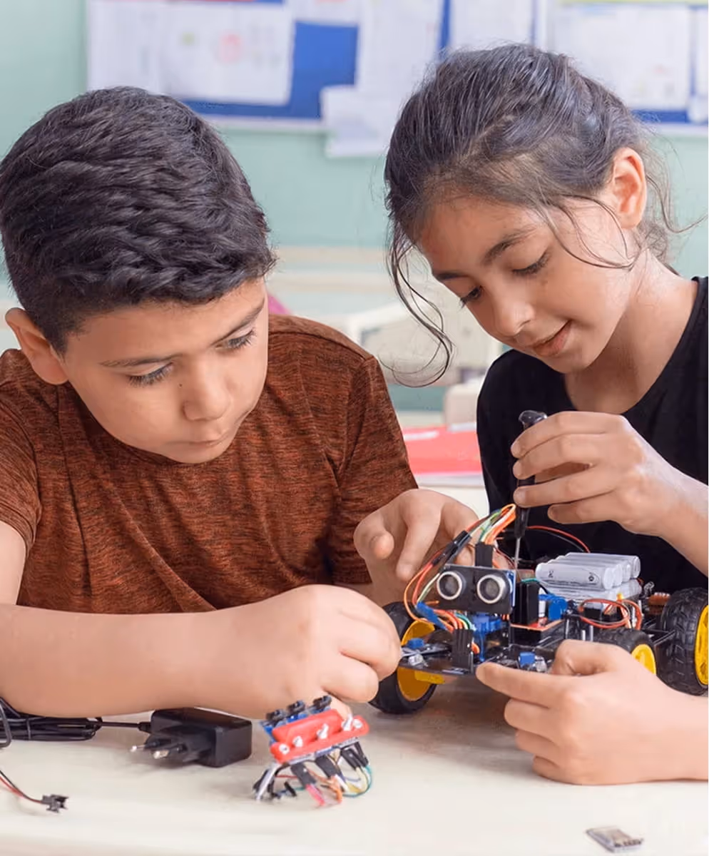 Two children assembling an electronic robot car with wires and circuits on a table.