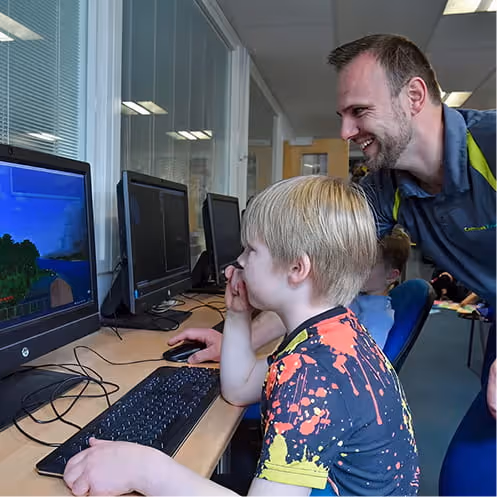 A teacher helps two boys use a laptop next to a table with a maze-like educational toy.