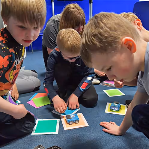 Children sitting on the floor engaging in a robotics activity with small blue robot cars and colored square cards.