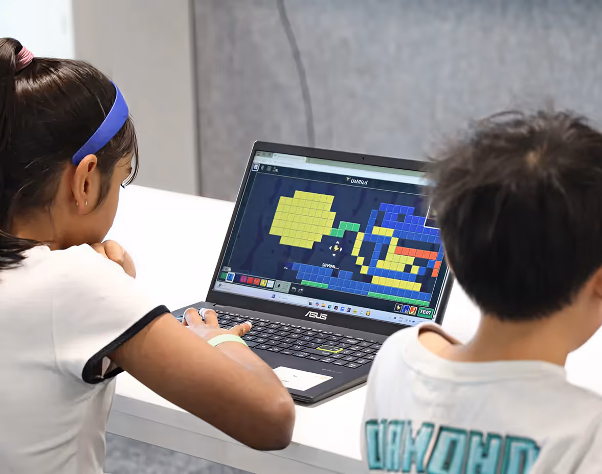 Adult male in a blue Computer Xplorers shirt helping a young boy in a red sweater with a laptop.