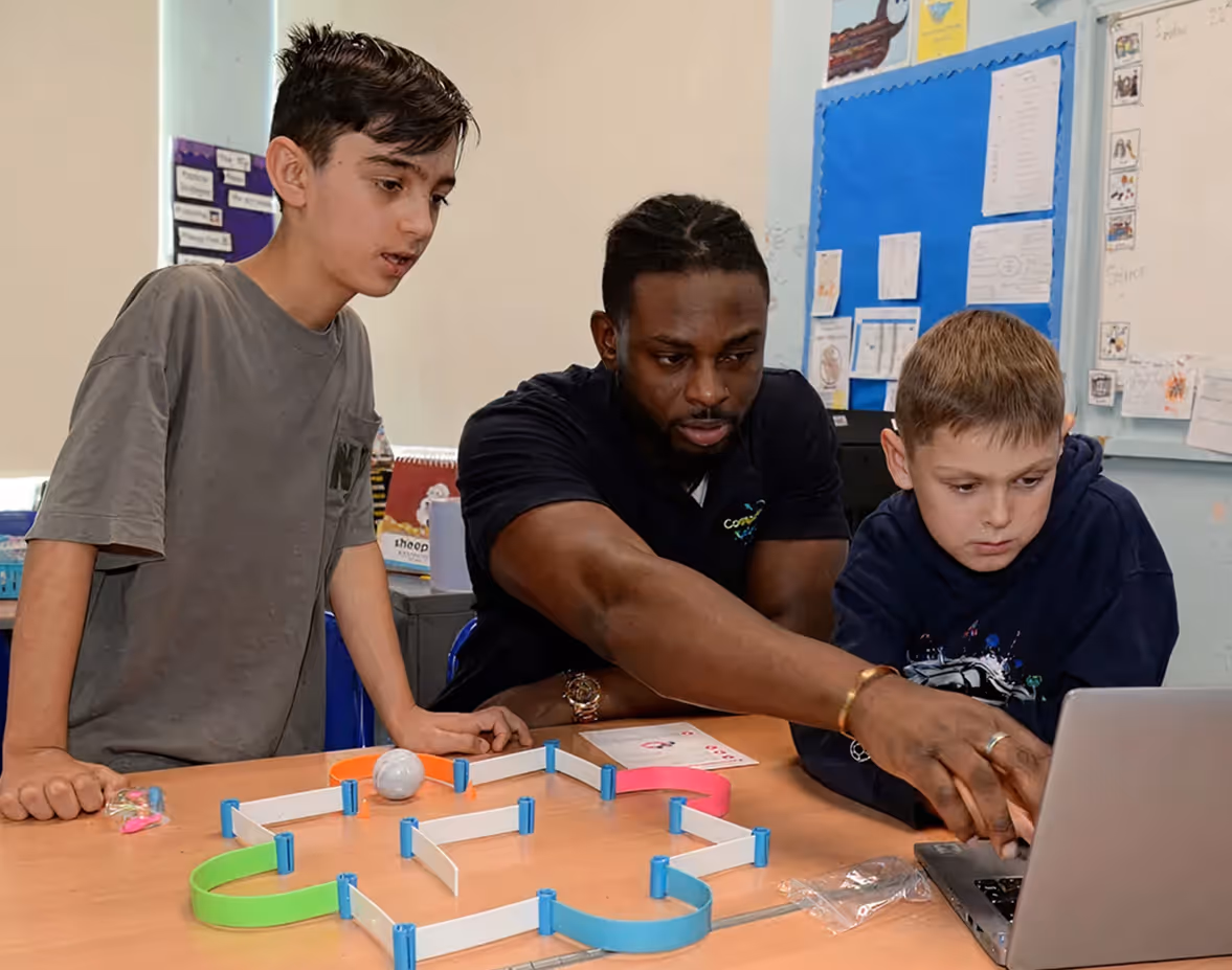 Instructor in a blue Computer Xplorers shirt helping a young boy in a red shirt with a laptop in a classroom.