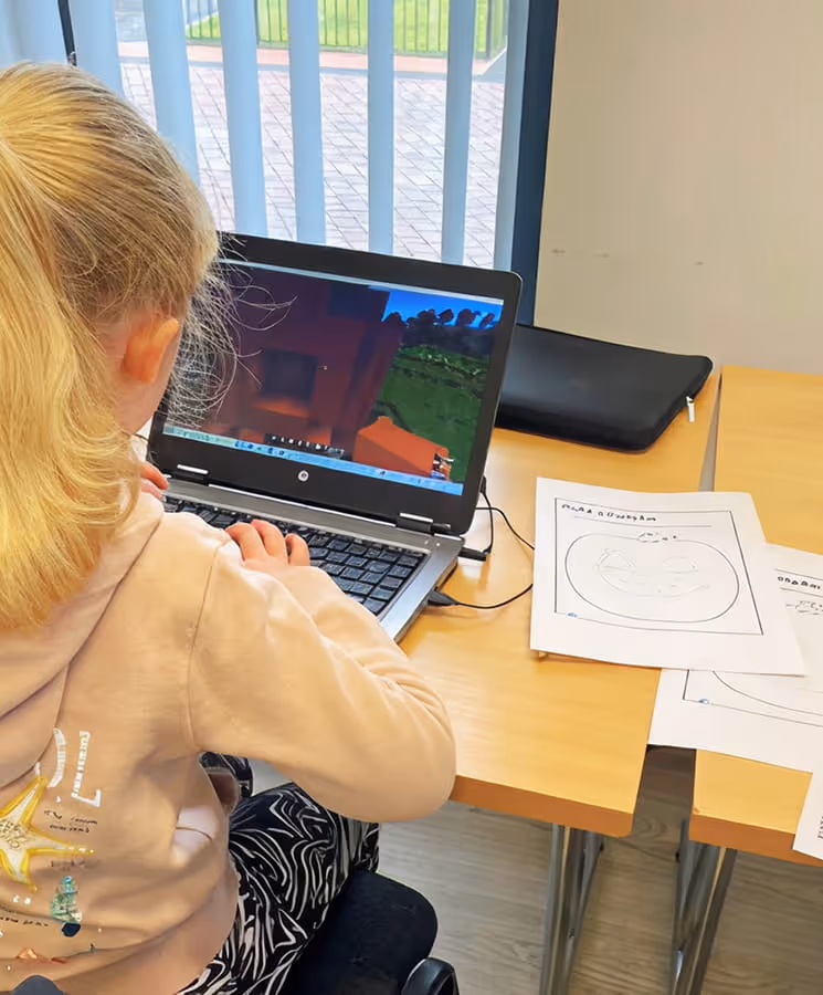 Child with blonde hair in a ponytail using a laptop computer at a desk with papers and a pencil case nearby.