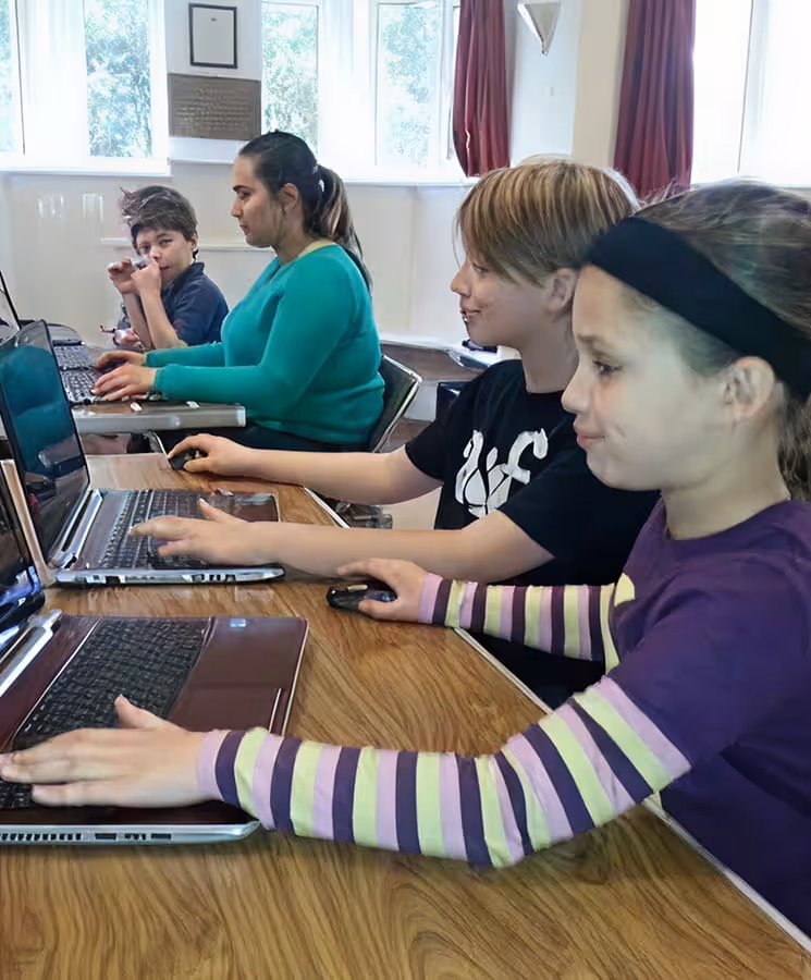 Four children sitting side by side at a wooden table, using laptops in a bright room with red curtains and large windows.
